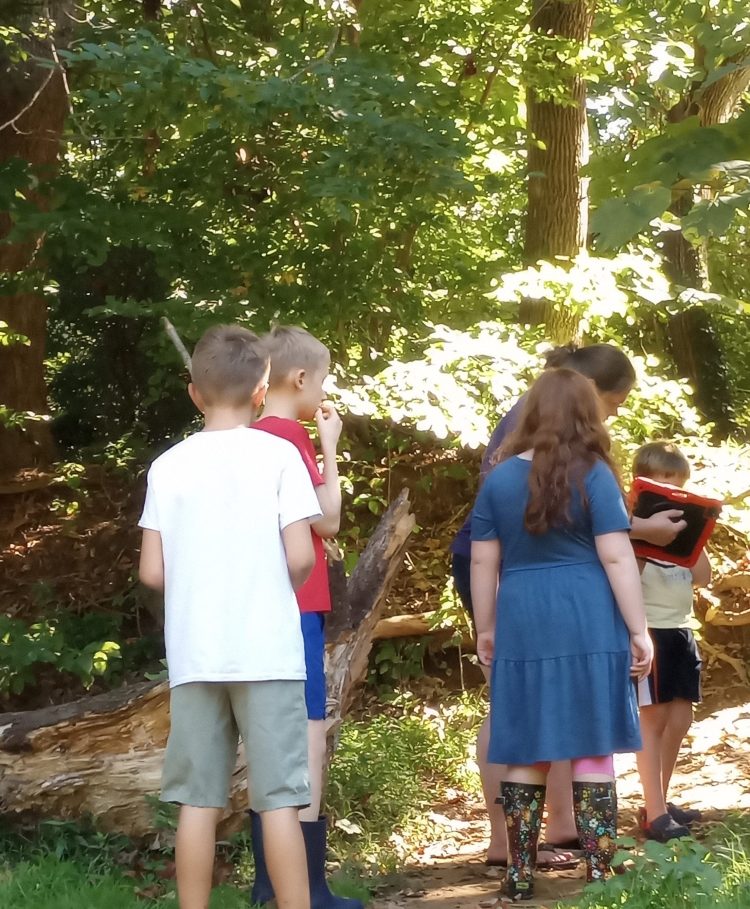 Children in a wooded area having a conversation. A woman holds an AAC device for one child to use to speak.